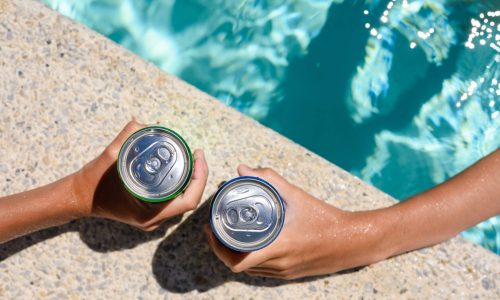 Two hands hold cans of cold drink in the pool on a hot day. The concept of vacation in a friendly company. Top view, selective focus on the bank.